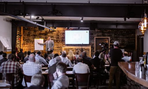A man stands on a well-lit stage, presenting processes for hiring BDMs with a digital whiteboard and a large scratch pad in front of a crowd.