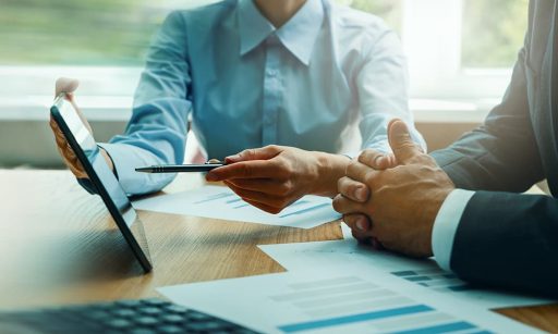Two business people sit down at a desk to negotiate with papers and a tablet.
