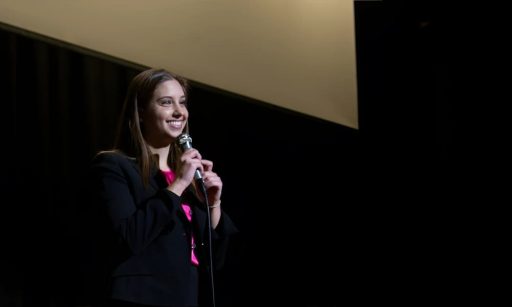 Julia Wojnar stands on a stage at a conference speaking into a microphone to a crowd of people.