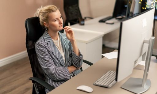 A disengaged employee sits at her desk in her office, working with a white monitor.