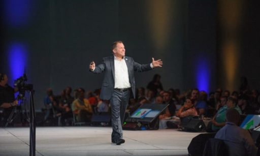 John DiJulius walks across a stage at a conference, speaking to a large crowd of people.